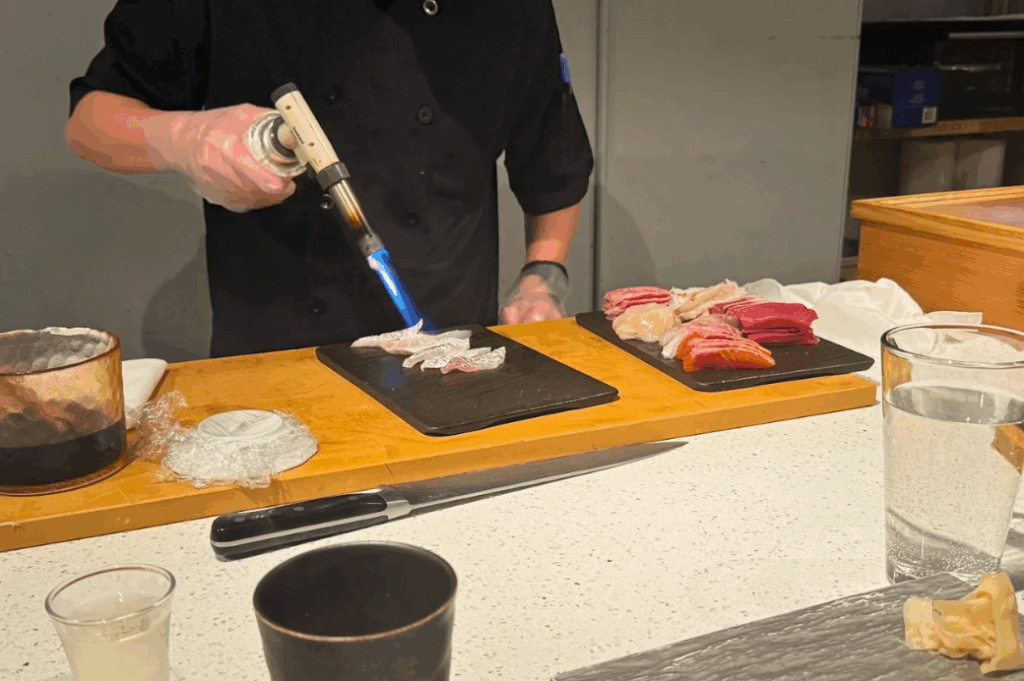 A SourAji chef using a blowtorch to sear a piece of fish on a black slate board at the counter