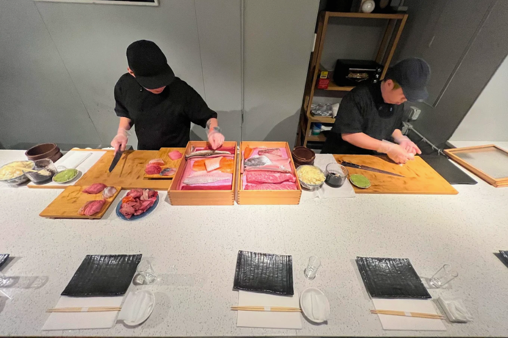 Two SourAji chefs slicing fish behind the sushi counter with boxes of tuna and wagyu laid out